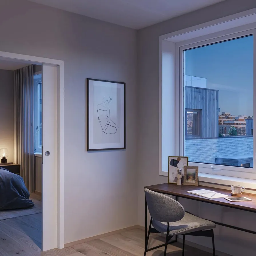 Arbeidsplass ved vindu med utsikt over taklandskap, og soverom synlig gjennom åpen dør.