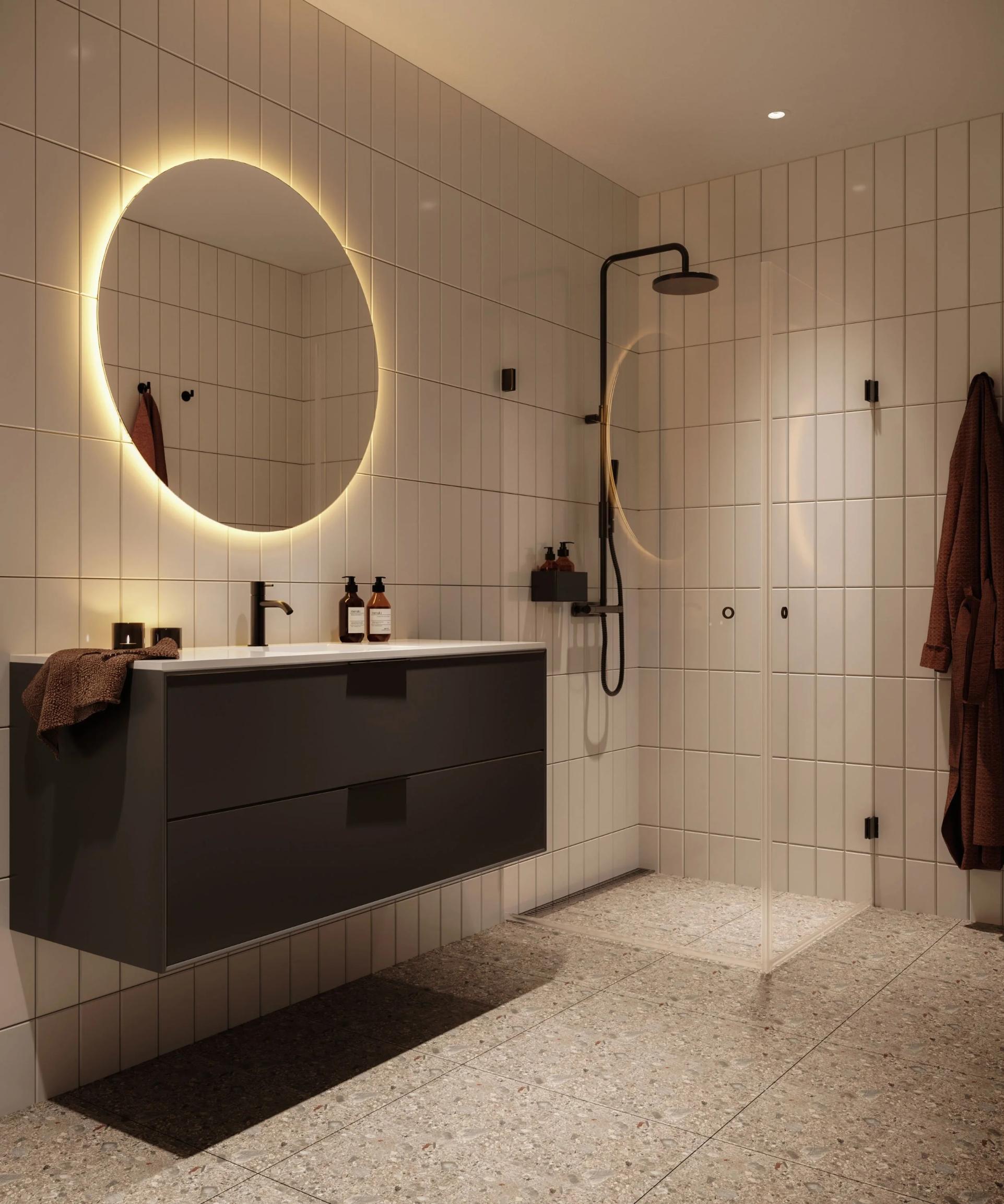 Bad med store fliser, rund bakbelyst speil, mørk servant og dusj med sorte detaljer.